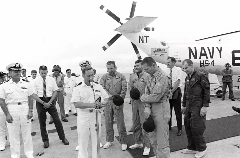 Bir gemi güvertesinde, çevresinde denizciler ve görevliler bulunan üç astronot