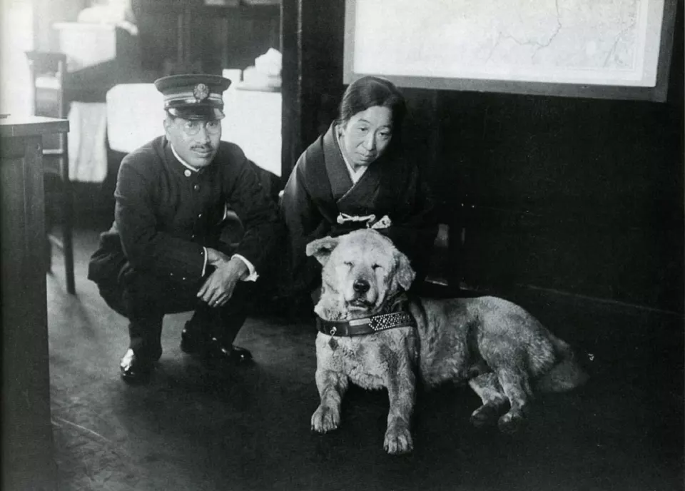 Bir istasyon binasının önünde, üniformalı bir adamın yanında duran beyaz tüylü, iri bir köpek