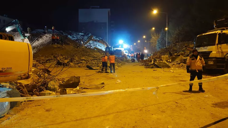 Gece ışıklandırılmış bir enkaz alanında turuncu yelekli kurtarma görevlileri ve iş makineleri