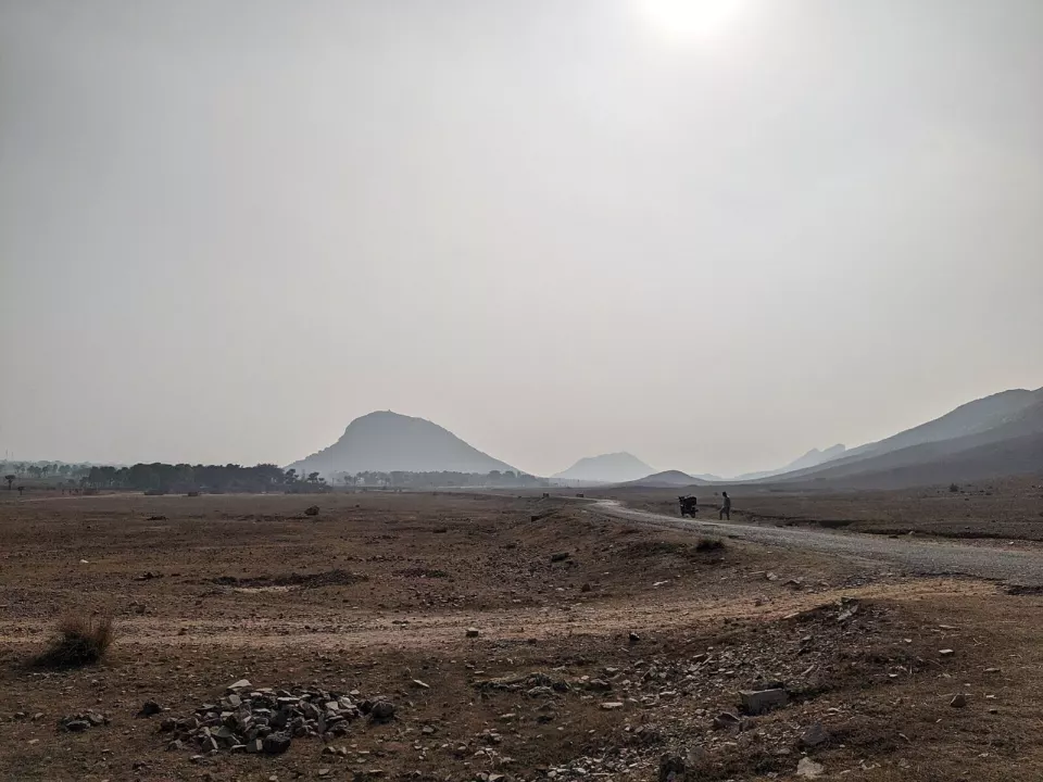 Puslu bir gökyüzü altında kuru, taşlı bir ova; uzakta sivri tepeler ve ovadan geçen toprak bir yol