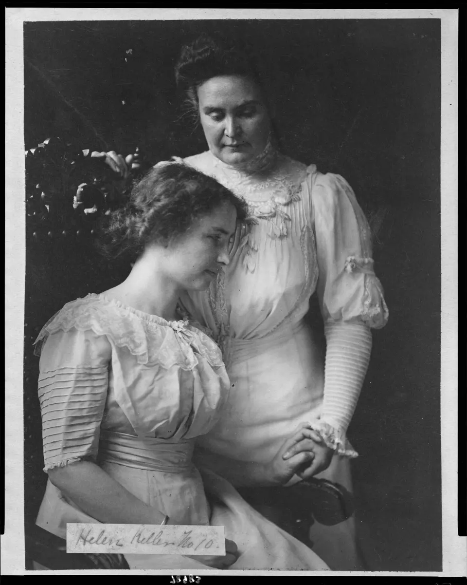 Oturan Helen Keller, yanındaki Anne Sullivan'ın elini tutuyor