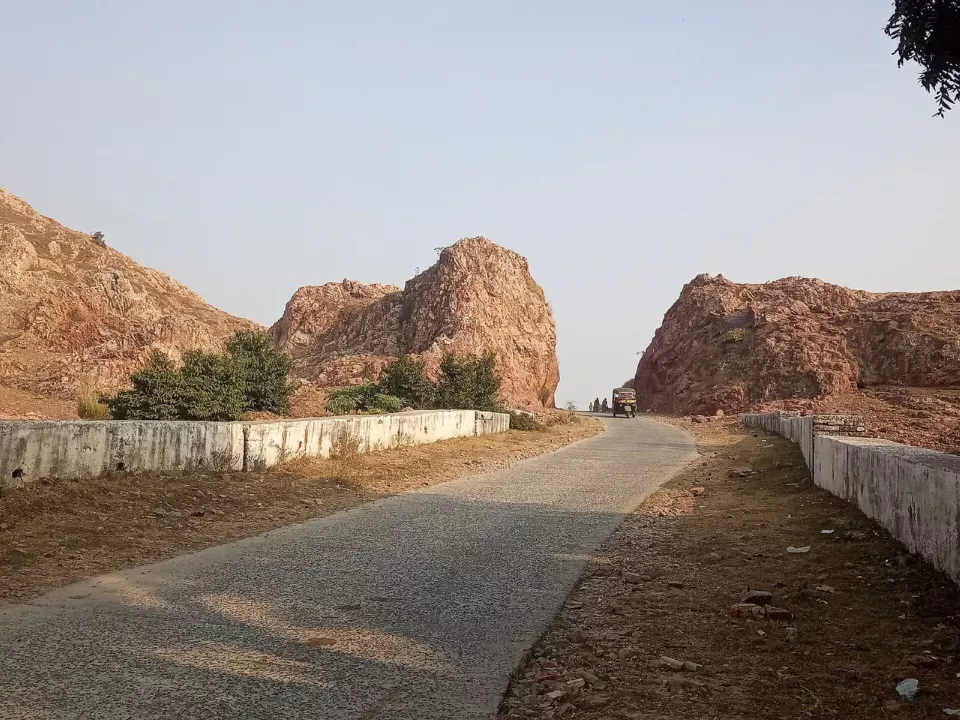 İki yanı kızıl kayalarla çevrili bir geçitten geçen, kenarları alçak duvarlı dar bir beton yol; uzakta bir üç tekerlekli araç