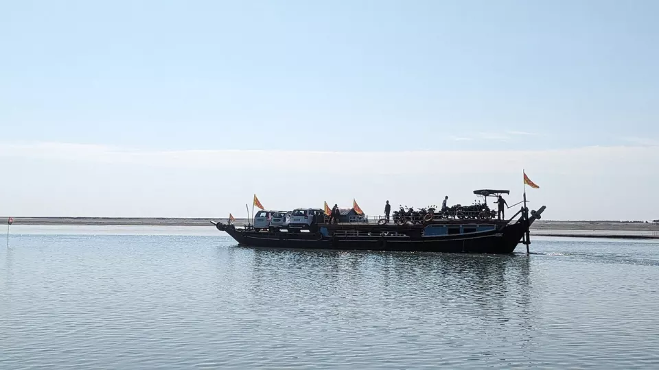 Durgun, geniş bir nehirde üzerinde araçlar ve insanlar bulunan, bayraklı uzun ahşap bir vapur; ufukta alçak kumluk kıyı